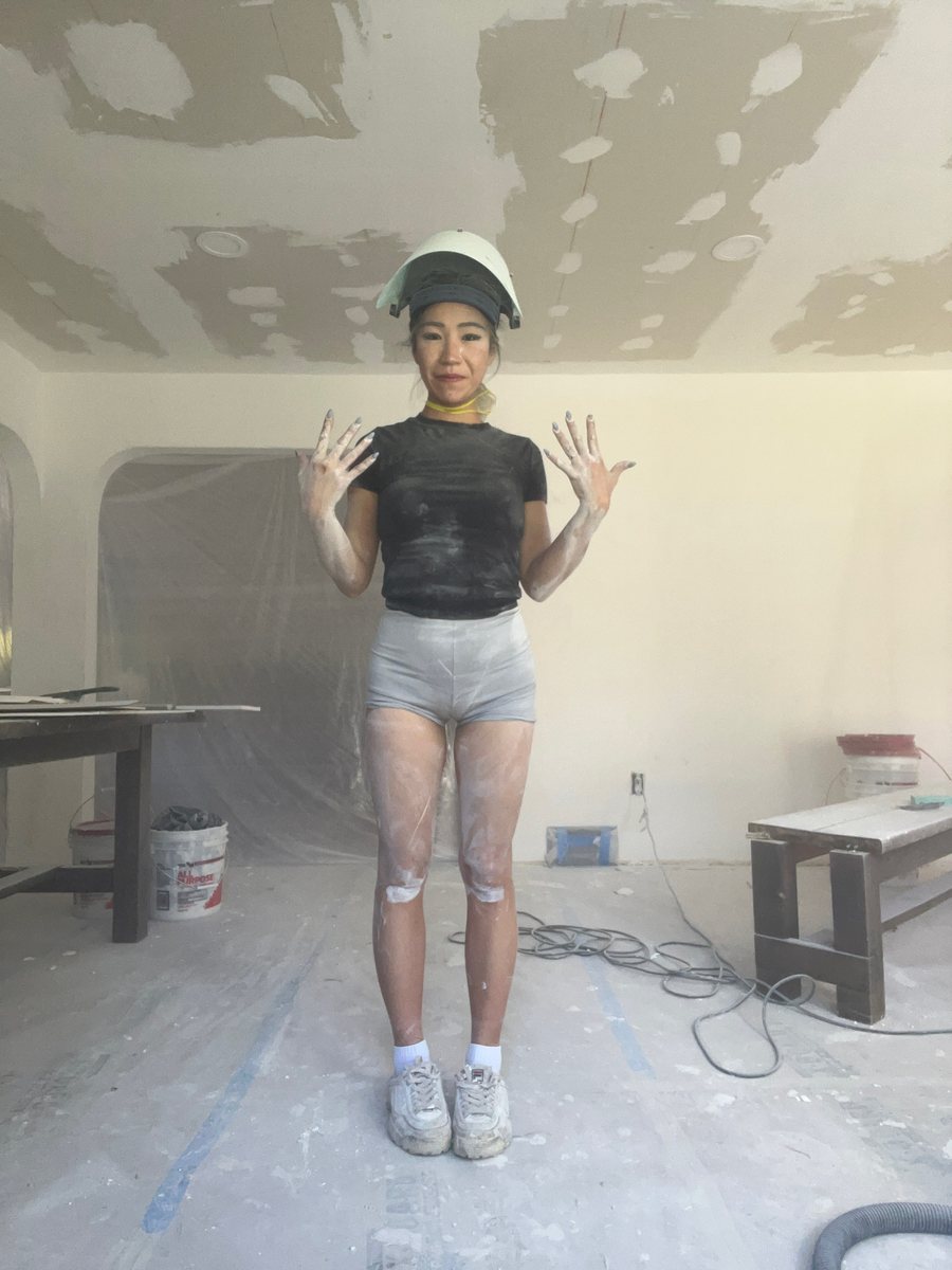 Everest Laina in a house under renovation, wearing a face shield, her hands and legs covered in drywall dust