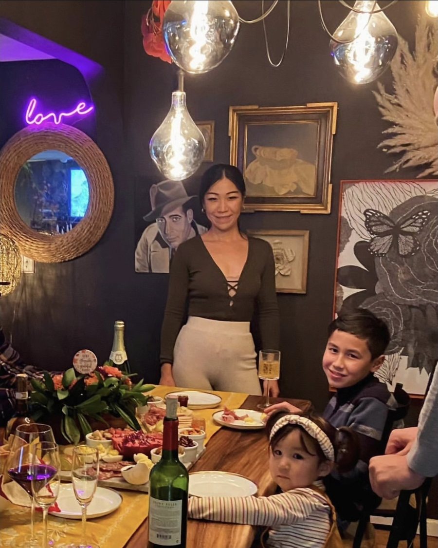 Everest Laina standing at the head of a holiday table with her children, in a dark room lit by glass pendant bulbs
