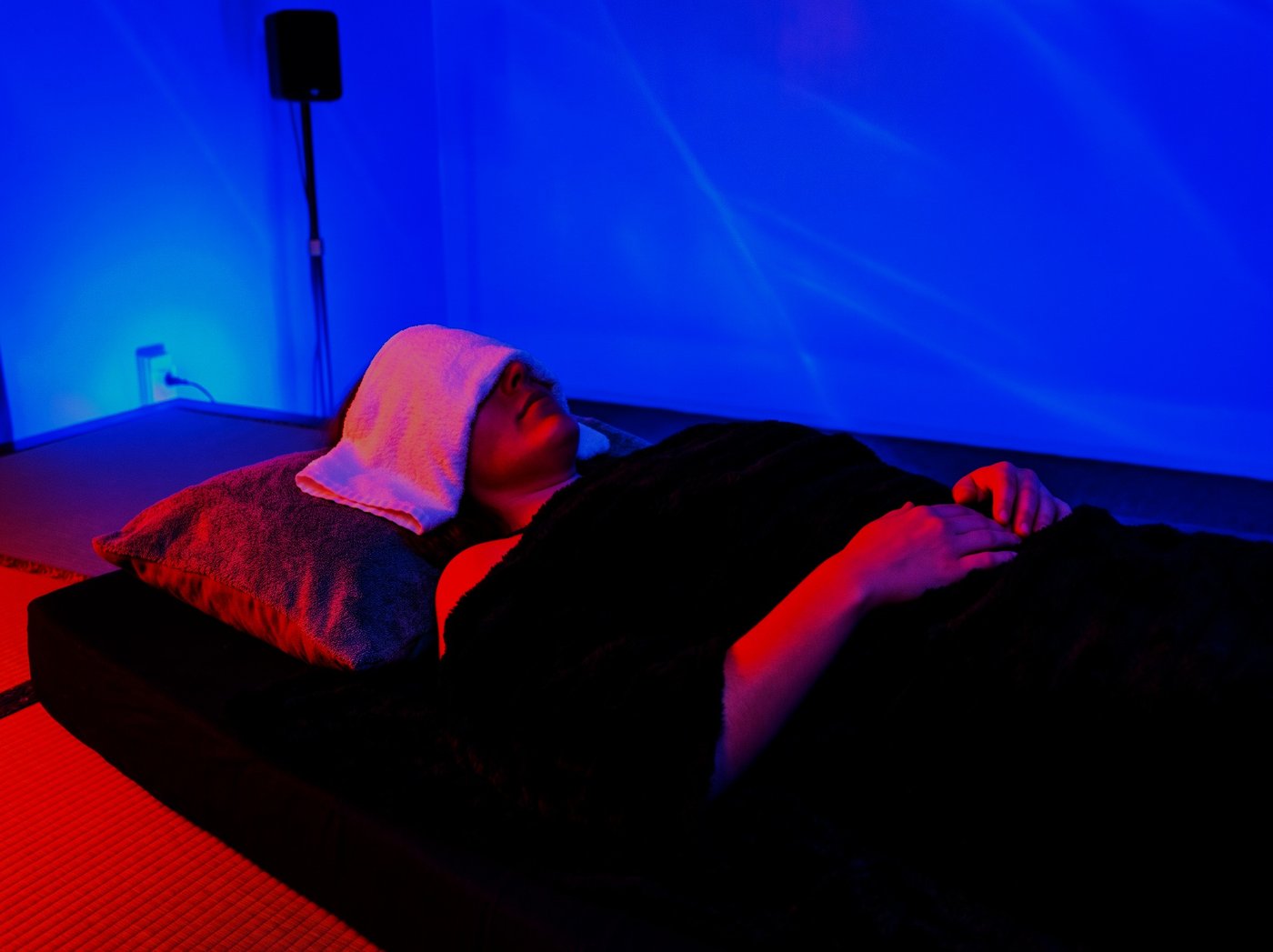 A person resting on the vibroacoustic platform under moving blue light, a warm towel over the eyes